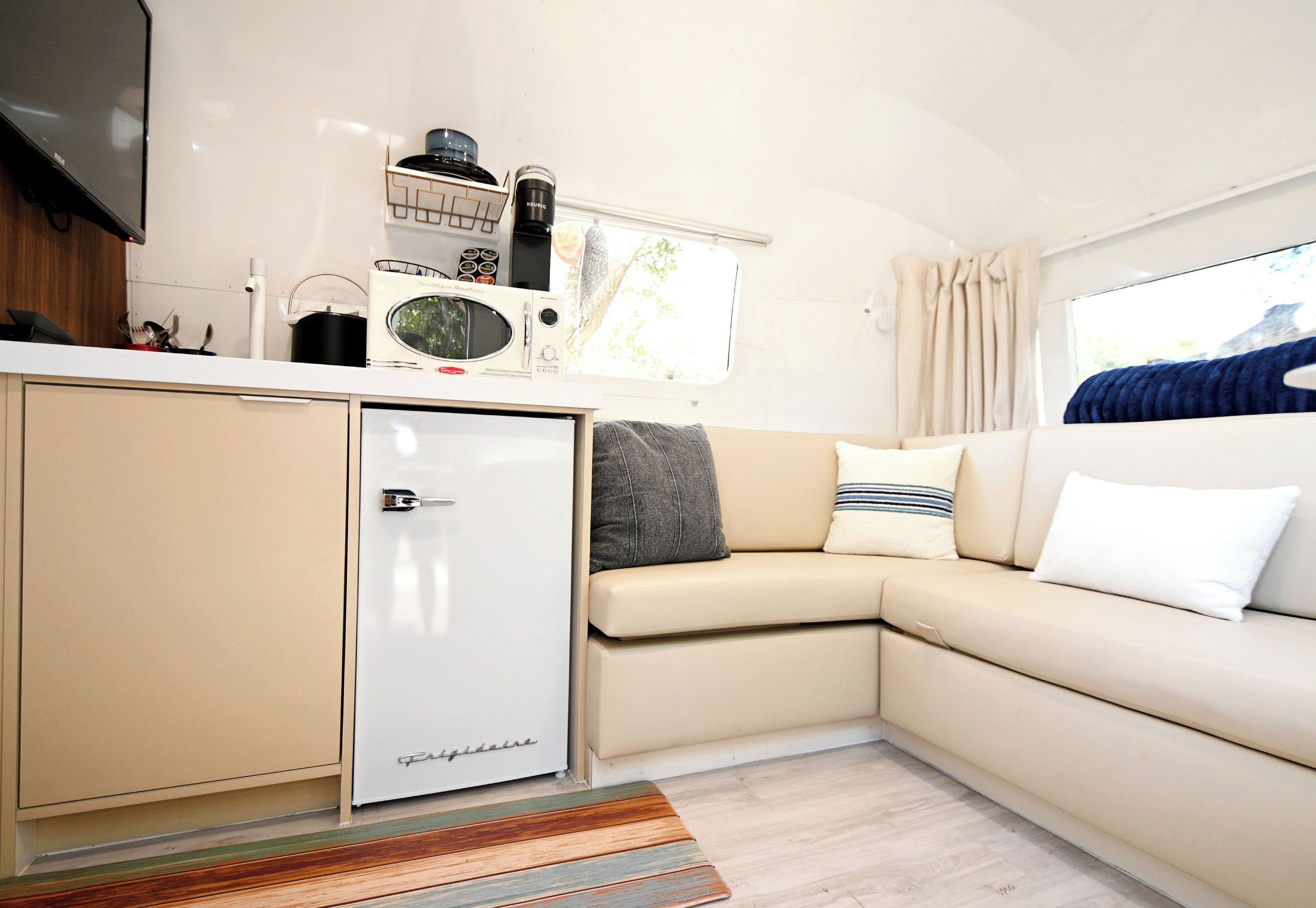 A compact kitchenette with a refrigerator, microwave, and coffee maker, a beige sofa with pillows, and a window with curtains.