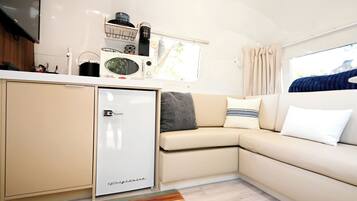 A compact kitchenette with a refrigerator, microwave, and coffee maker, a beige sofa with pillows, and a window with curtains.