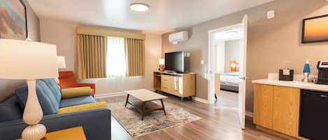 A modern hotel room with a sofa, a small coffee table, a TV, a kitchenette, and a bathroom visible through an open door.