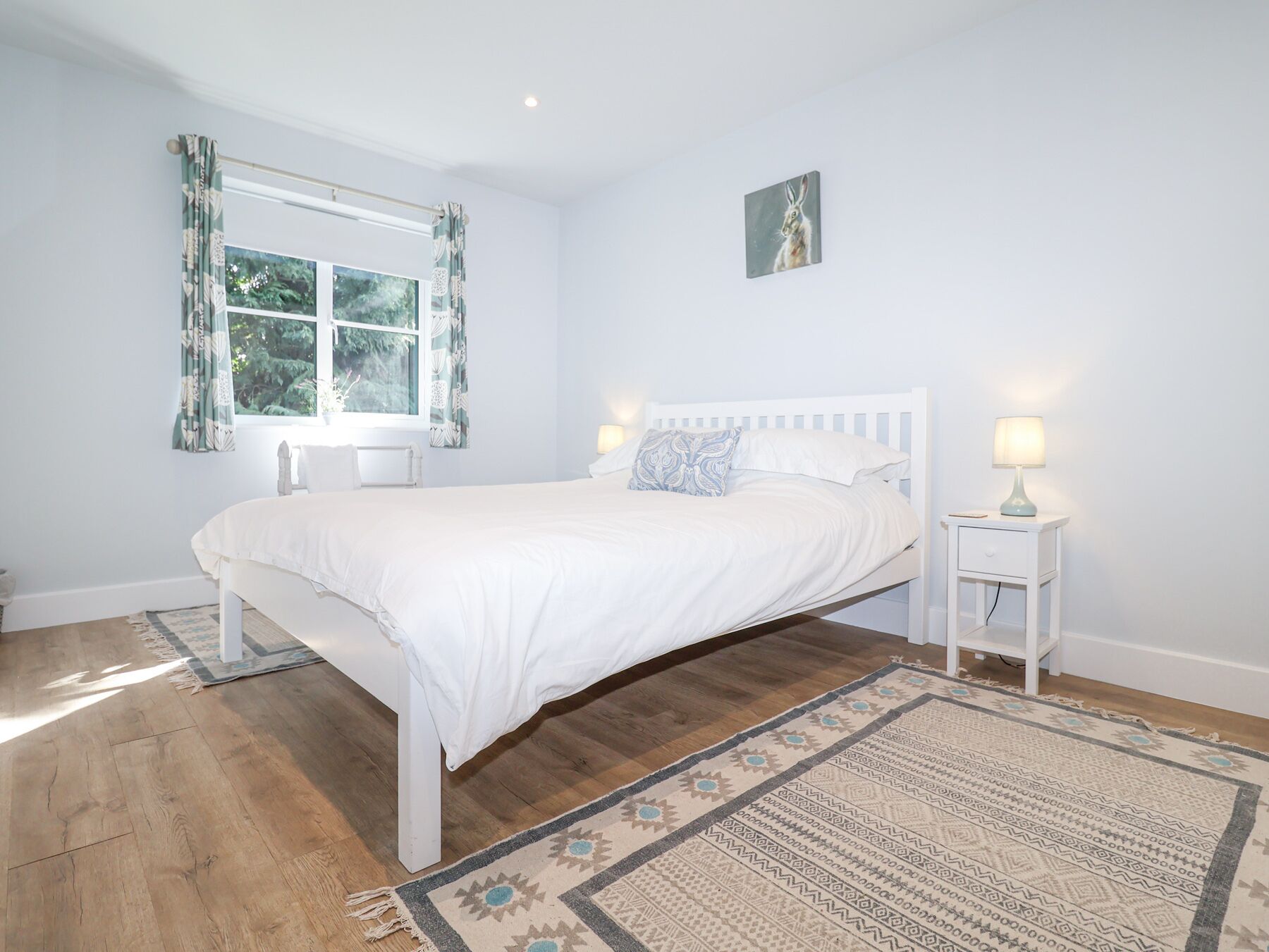 A bedroom with a bed, two bedside tables with lamps, a window with curtains, a rug, and a painting on the wall.