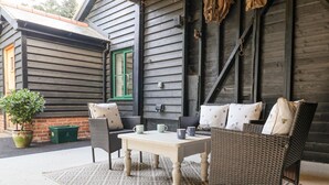 A cozy outdoor seating area with wicker furniture and a table, set against a wooden building with a green window. - Bradley's Barn (Colchester)