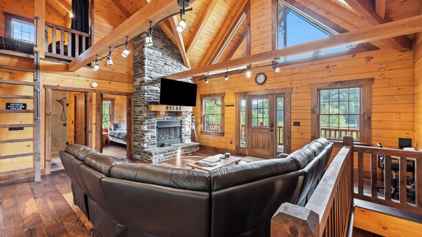 A cozy cabin interior with a stone fireplace, leather seating, wooden beams, and large windows.
