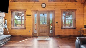 A wooden cabin interior with a large wooden door, two windows, a clock, and a stone fireplace.