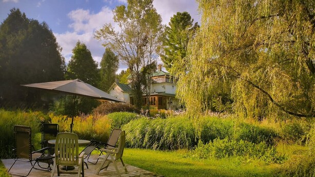 An outdoor patio with a table and chairs, surrounded by lush greenery and a house with a green roof in the background. - Vintage Inspiration & Creative Tools in a Victorian Manor (Namur)