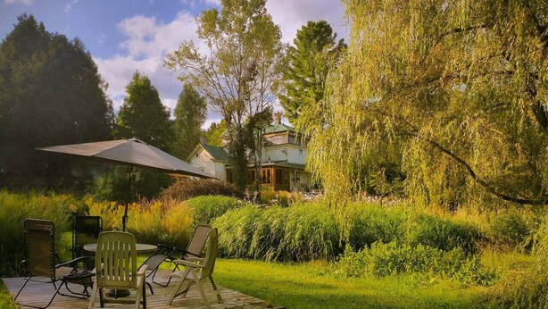 A patio with a table and chairs, surrounded by lush greenery and a house in the background. - The Cinema Room with Spa - Le Salon des Inconnus (Namur)