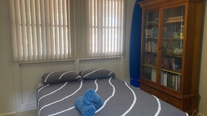 Une chambre à coucher avec un lit, des oreillers, une serviette bleue, une fenêtre avec des stores et une bibliothèque en bois.