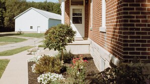 Une maison à deux étages, construite en briques, avec un porche à l’avant, un petit jardin agrémenté de diverses plantes et fleurs, et un garage blanc à l’arrière-plan.