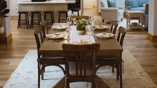 Un coin repas avec une grande table en bois, dressée pour six personnes, entourée de chaises en bois. Un lustre est suspendu au-dessus de la table, et la pièce est agrémentée de meubles blancs et d’un tapis à motifs.