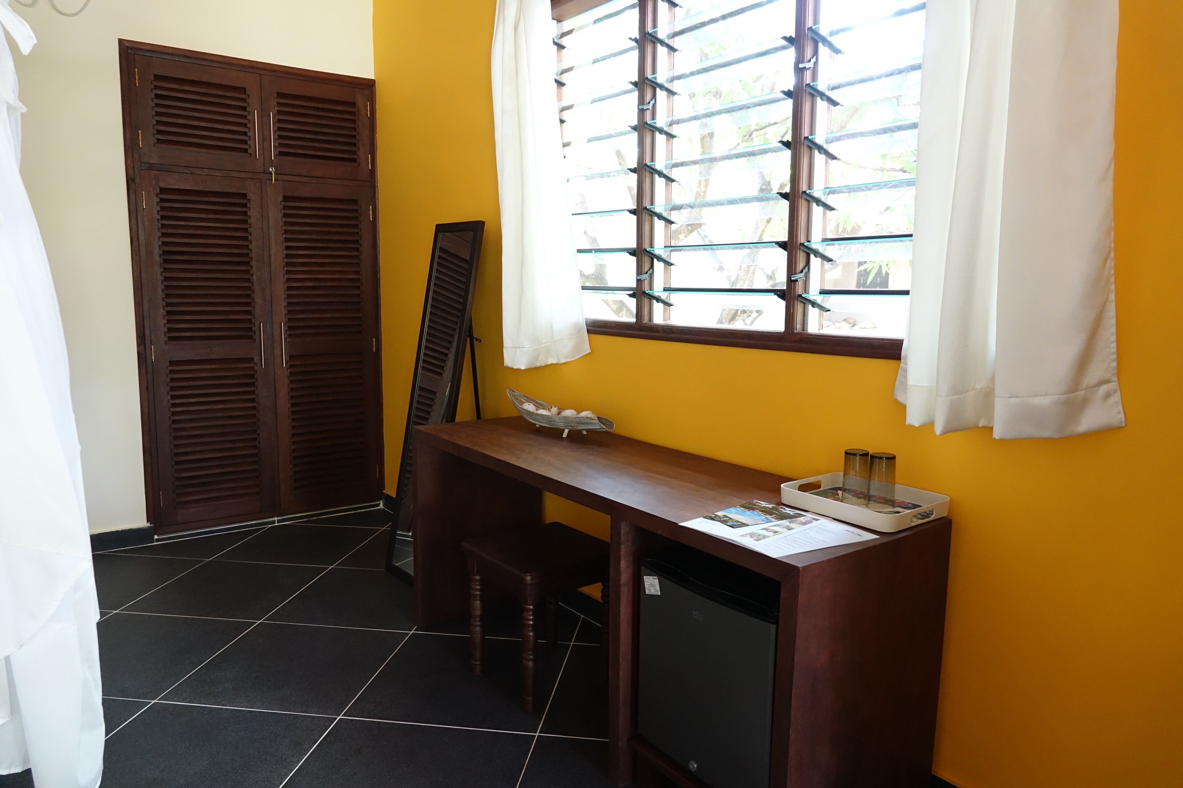 A room with a wooden desk, a small table, a TV, and a window with blinds.