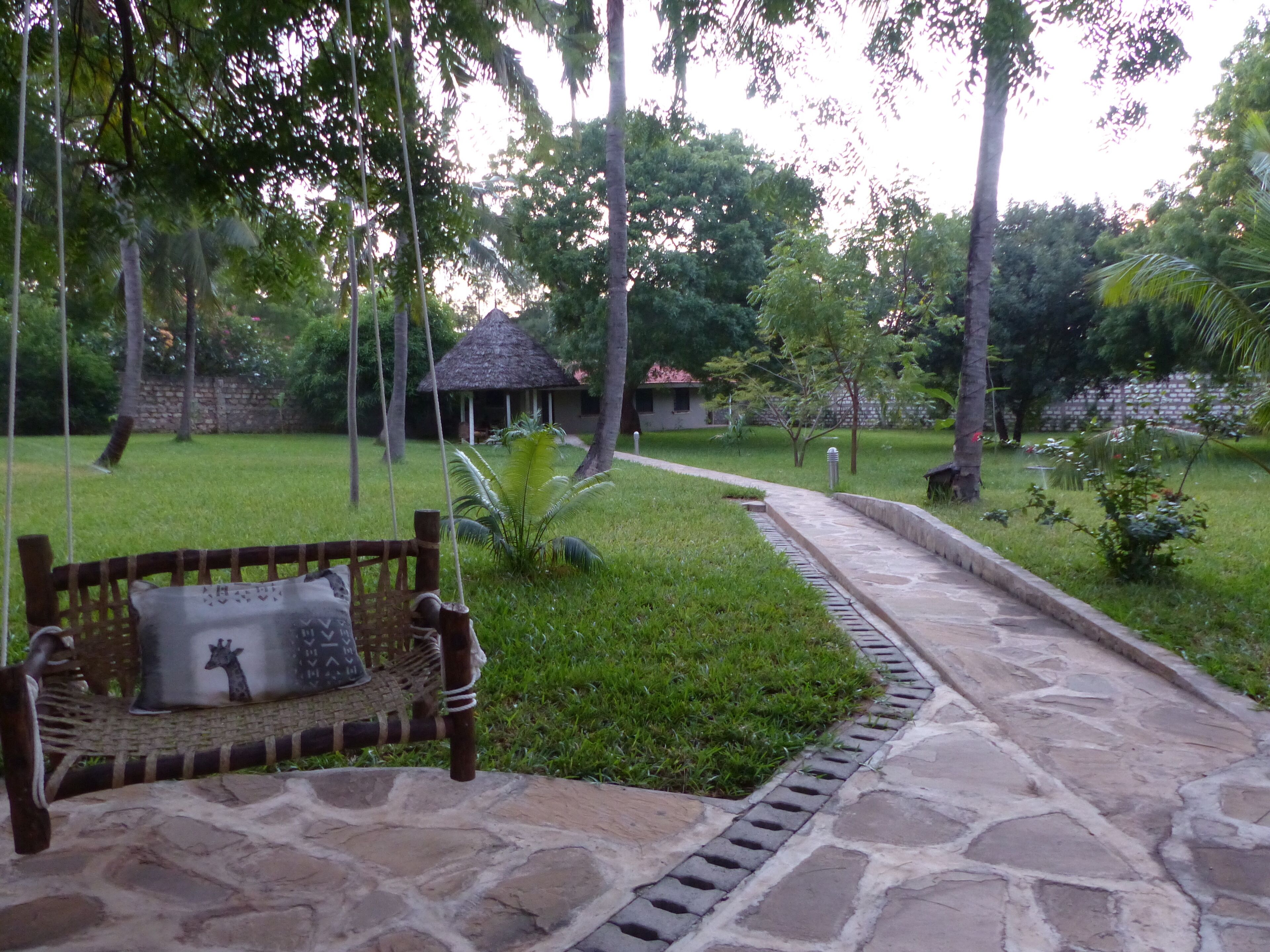 A stone pathway leading to a thatched-roof structure surrounded by lush greenery and tall trees.