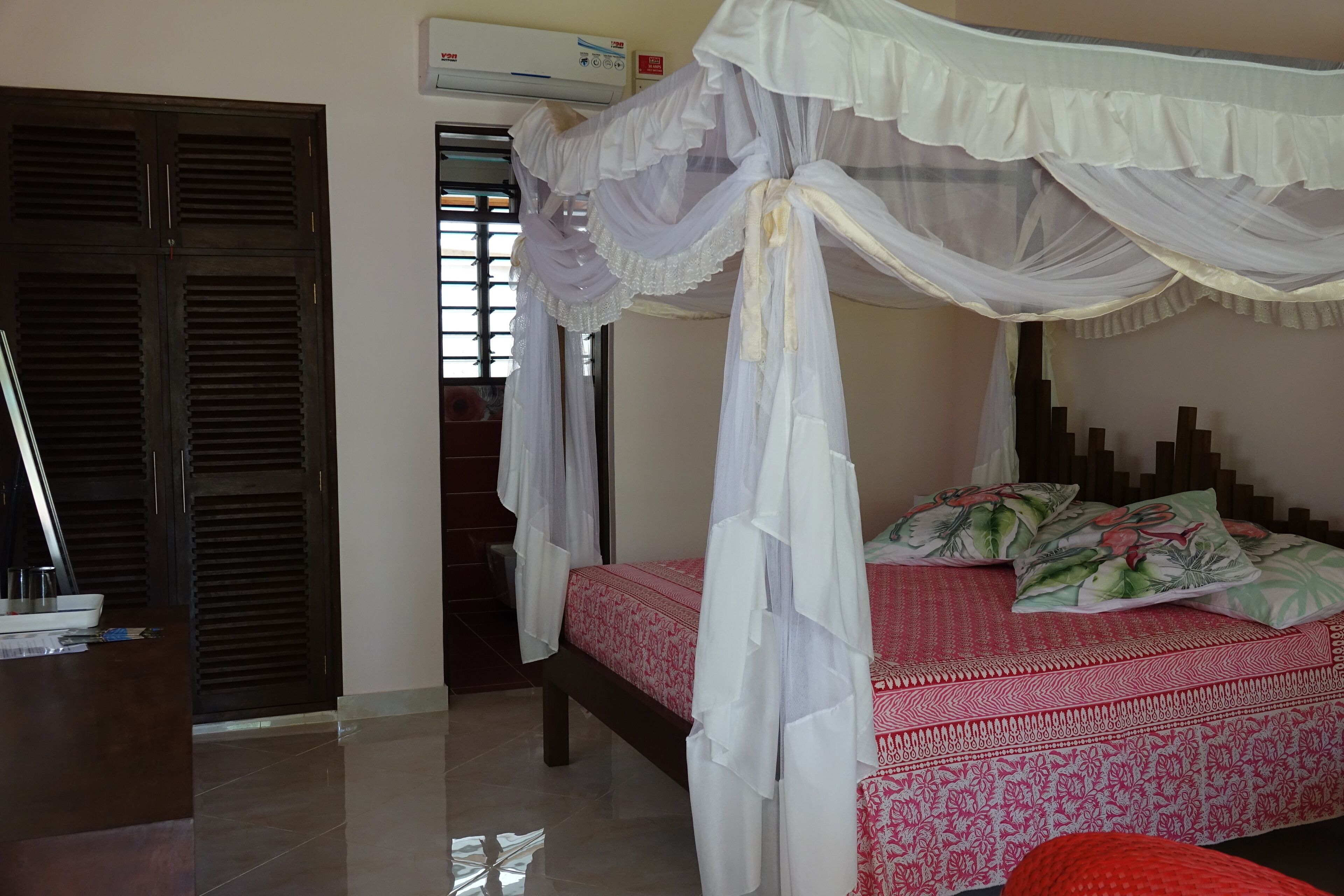 A bedroom with a bed under a canopy, a wooden wardrobe, and a window with blinds.