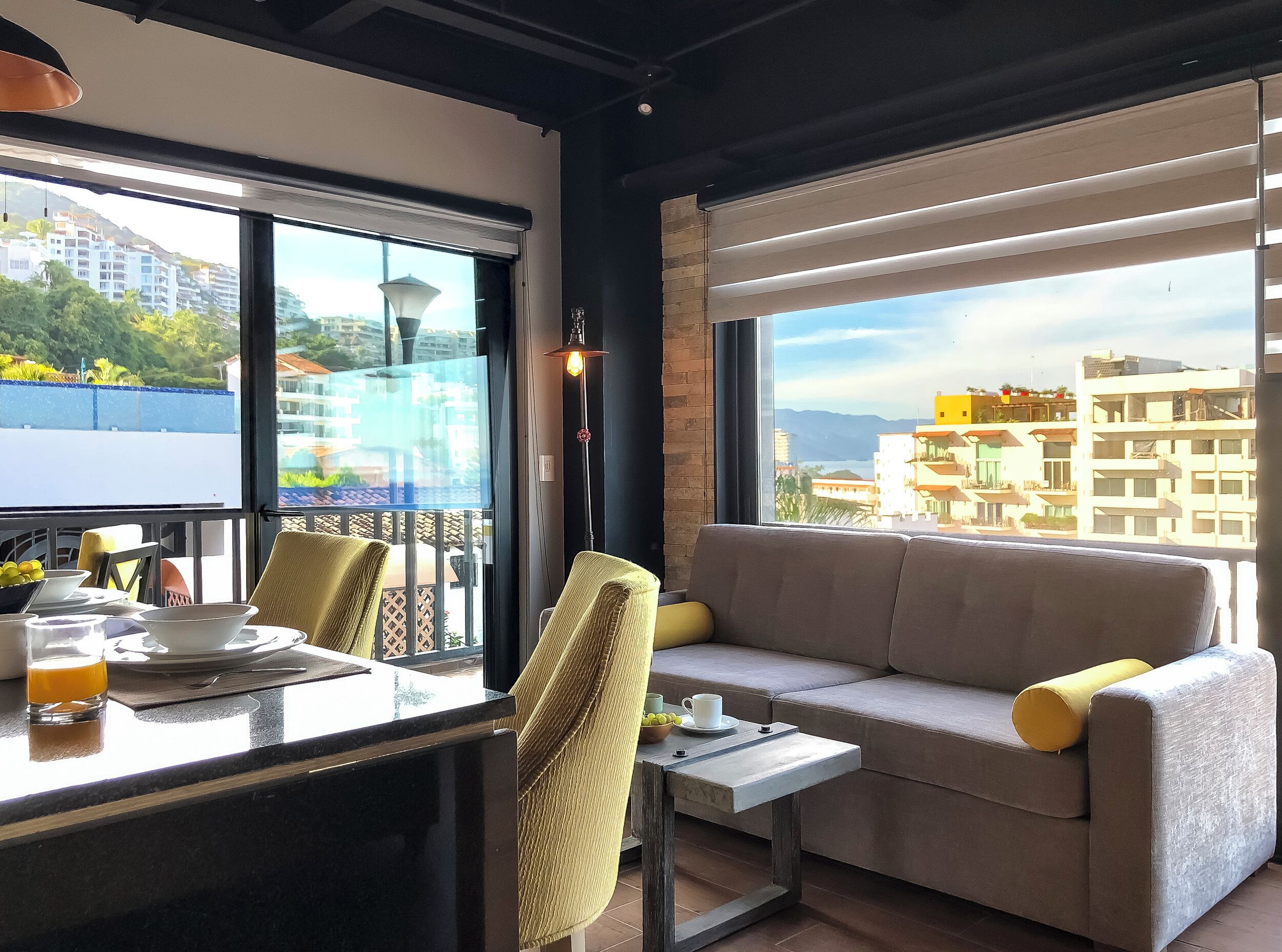 En moderne stue med sofa, stole, et lille bord og en balkon med udsigt over bygninger og grønne områder.