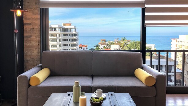 A modern living room with a grey sofa, a small table with a candle, a water bottle, and a bowl of grapes. A large window offers a view of the sea and buildings.