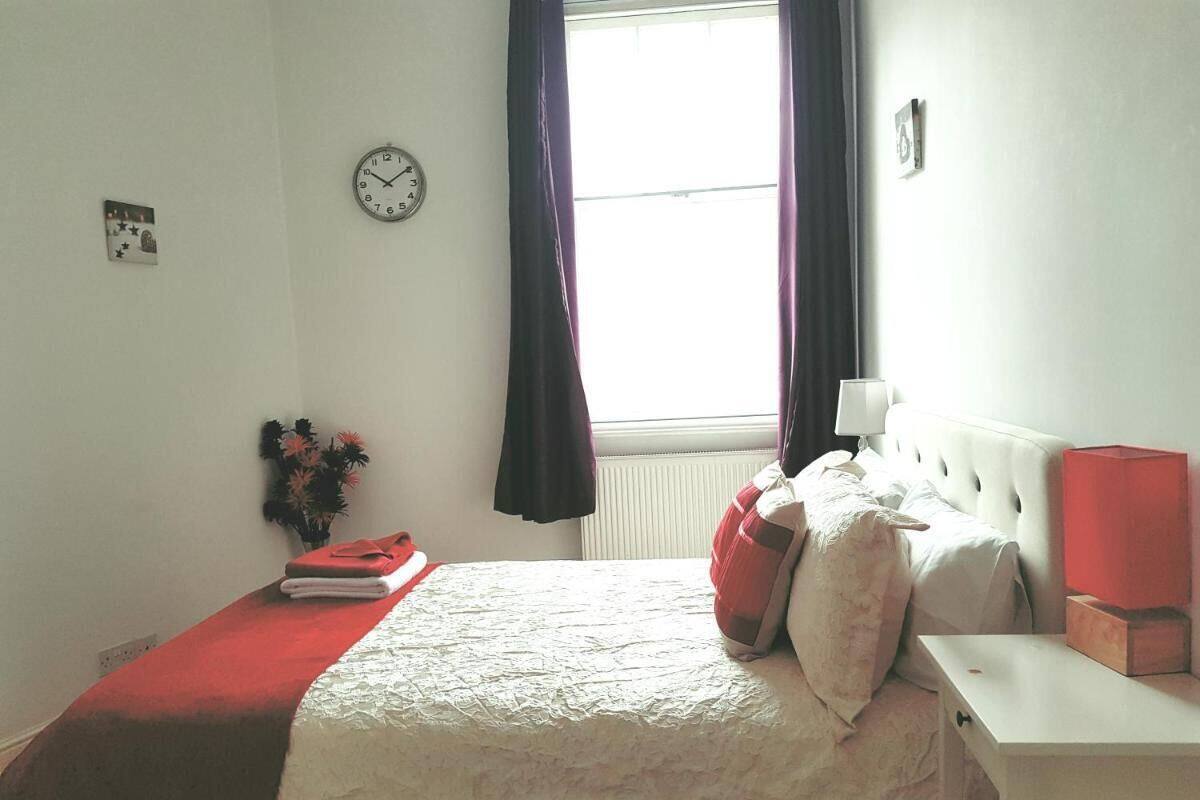 Un dormitorio con cama, mesita de noche, lámpara, reloj y ventana con cortinas.
