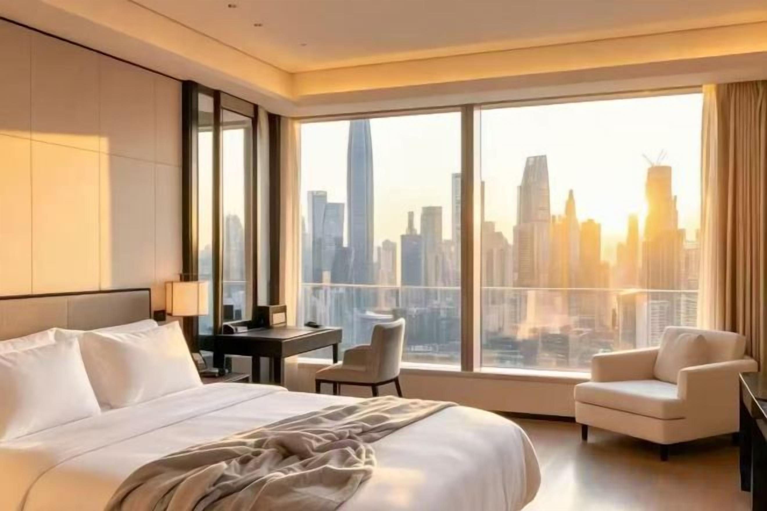 A hotel room with a large bed, a desk, a chair, and a view of a city skyline through large windows.