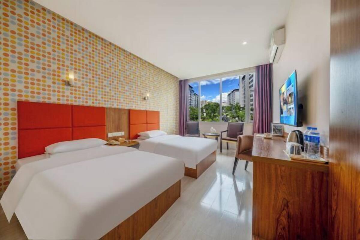 A hotel room with two beds, a wooden desk, a TV, and a window with a view of buildings.