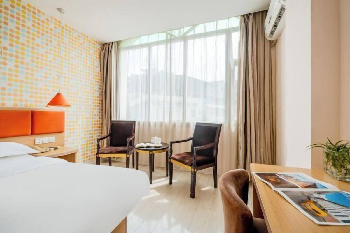 A hotel room with a bed, two chairs, a small table, a lamp, and a window with curtains.