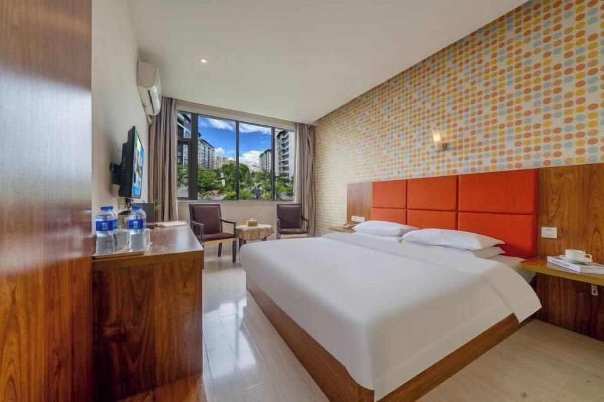A hotel room with a large bed, a desk with water bottles, a TV, and a view of buildings and trees.