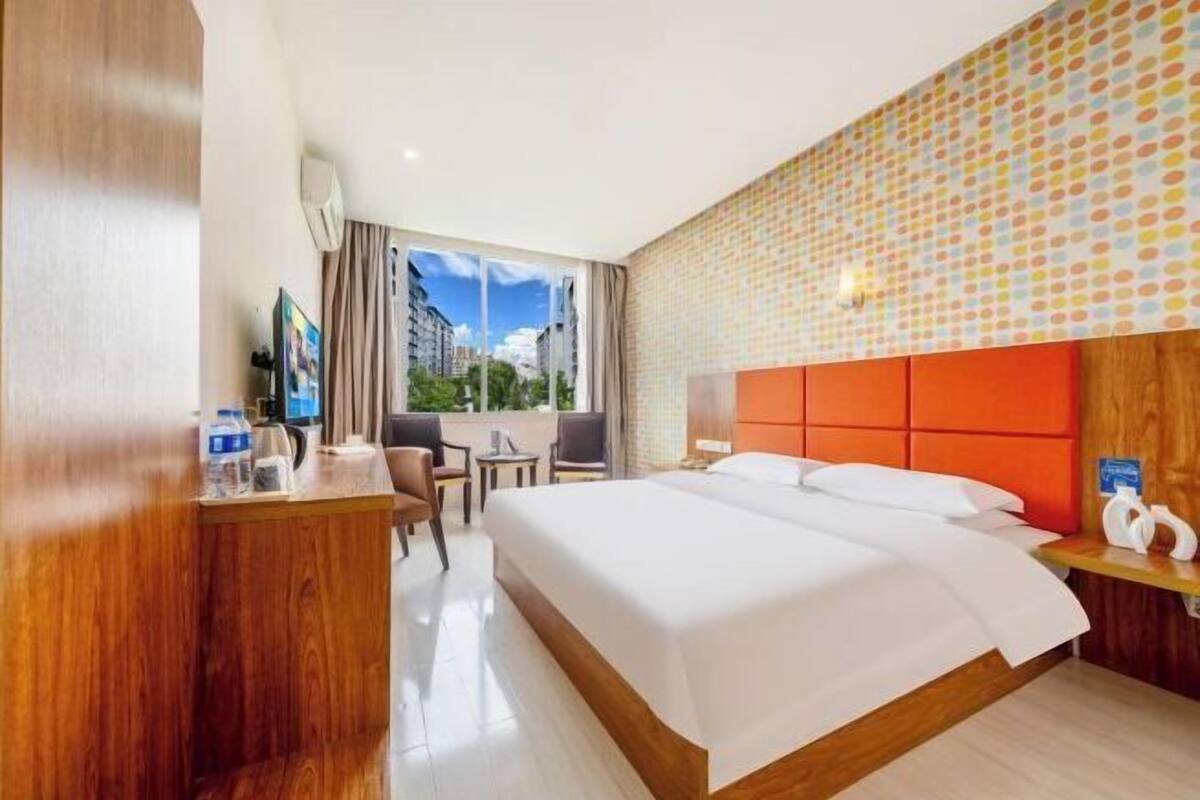 A hotel room with a large bed, a desk, a chair, a window with a view of buildings, and a patterned wall.