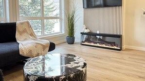 Una sala de estar moderna con un ventanal grande, un sofá negro, una mesa de centro de mármol, un televisor de pantalla plana montado en una pared inclinada y un pequeño acuario.