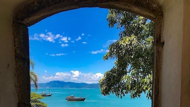 A view through an archway of a coastal area with boats, a pier, and people on the beach.