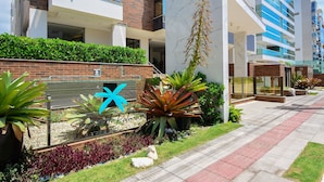 A modern building with a brick facade, balconies, and a landscaped entrance with plants and a sign.
