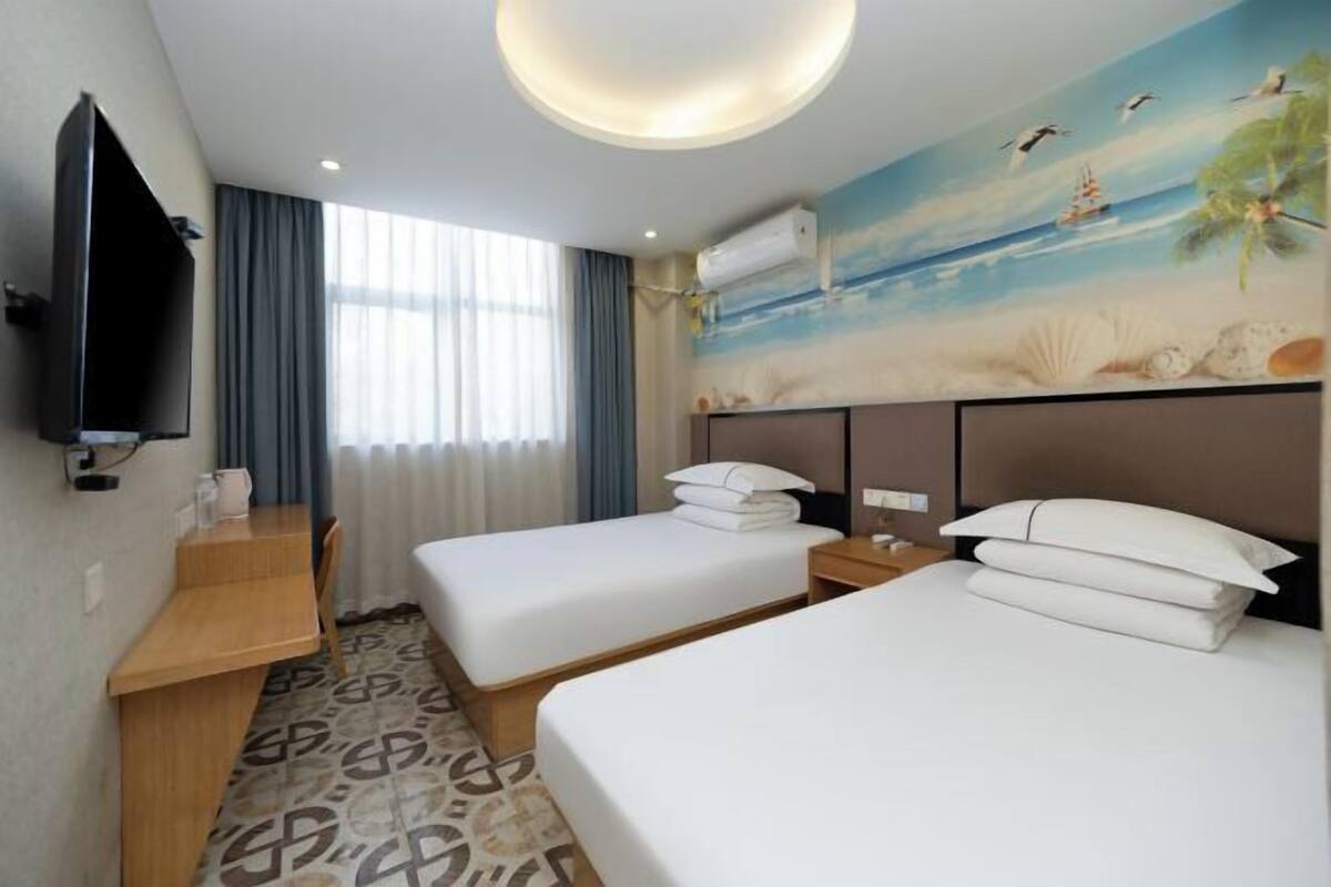 Ein Hotelzimmer mit zwei Betten, einem Schreibtisch, einem Fernseher und einem Wandbild mit einer Strandansicht.