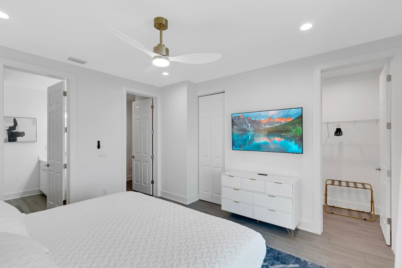 A bedroom with a large bed, a ceiling fan, a mounted TV, and a white dresser.