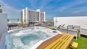 A hot tub with a wooden table and lounge chairs, overlooking a beach and a hotel building.