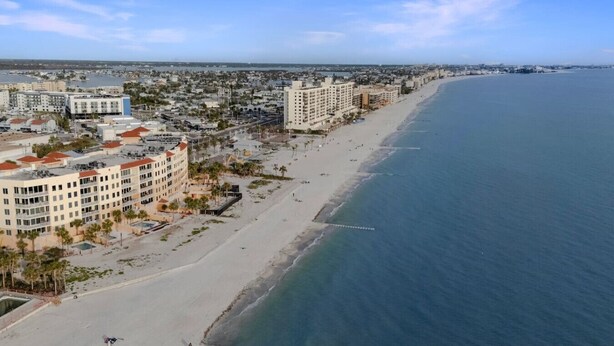 An aerial view of a coastal city with a long sandy beach, several multi-story buildings, and a clear blue ocean.