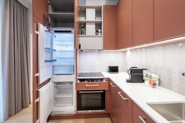 A compact kitchen with a built-in oven, a refrigerator, and a countertop with a coffee maker. - Designer Parkside Apartments (Riga)
