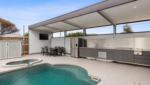 Une cuisine extérieure moderne avec une piscine, un espace repas et un téléviseur fixé au mur.