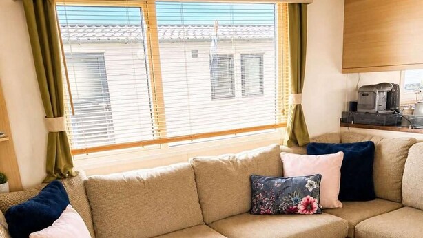 A cozy living room with a beige sectional sofa, a wooden coffee table, and a window with blinds. - Caravan Meg Sleeps 6 With Free Parking (Rhyl)