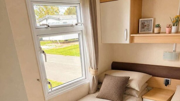A compact bedroom with a single bed, a small bedside table, a nightstand, a window with curtains, and a shelf with decorative items. - Caravan Meg Sleeps 6 With Free Parking (Rhyl)