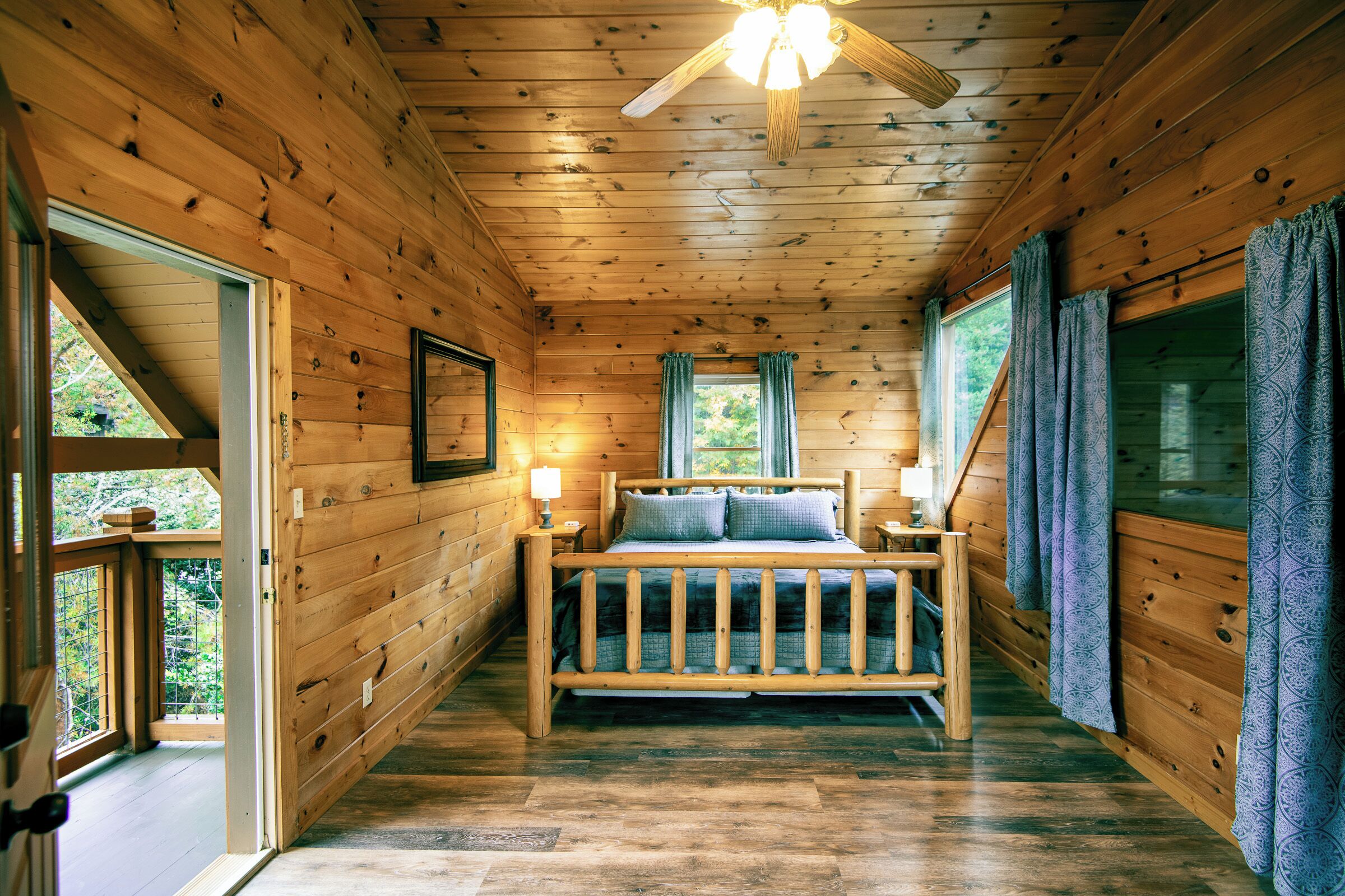 A wooden cabin bedroom with a bed, two windows, a ceiling fan, and a door leading outside.