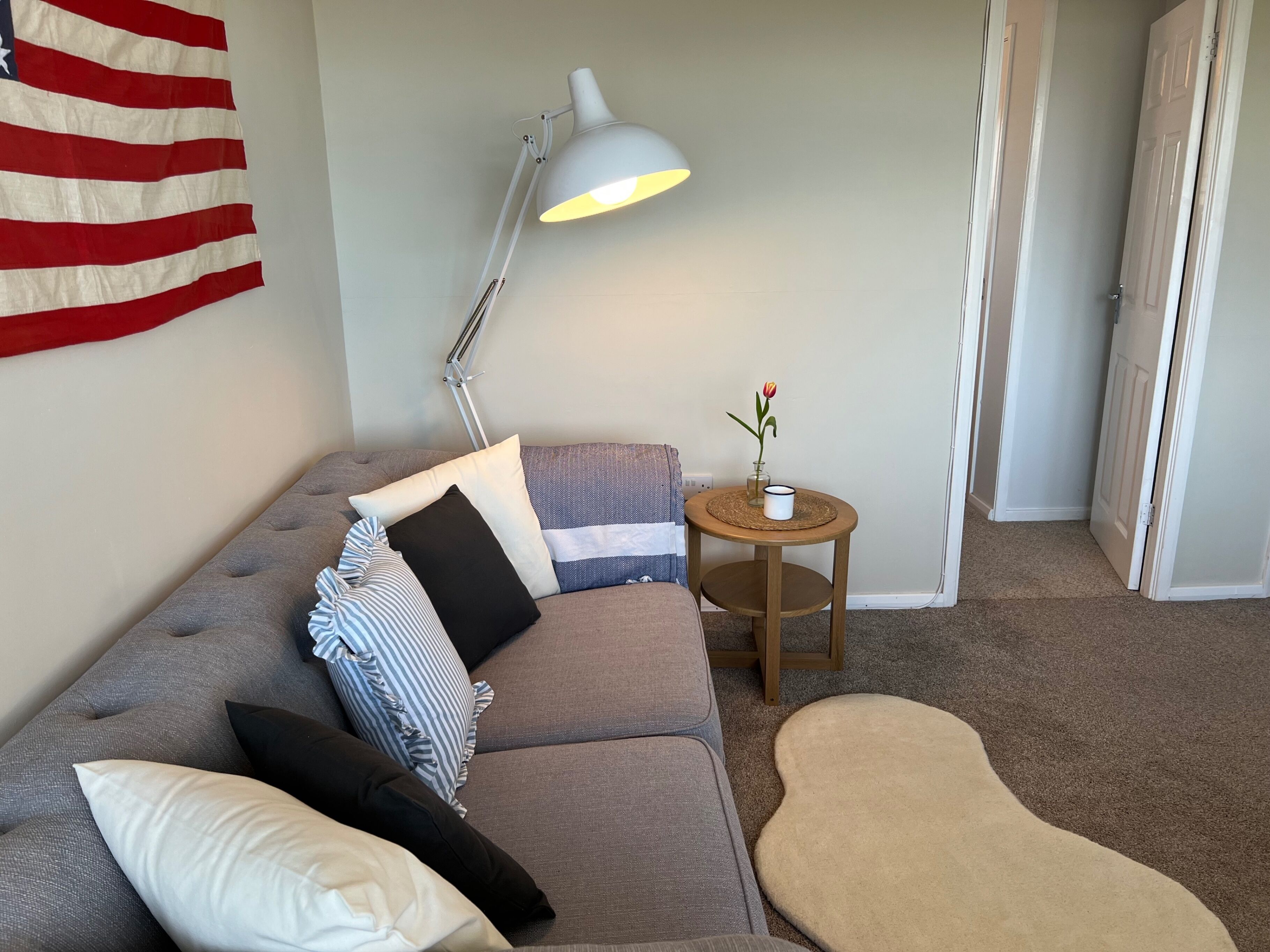 A living room with a grey sofa, a white lamp, a small wooden side table with a vase and mug, and a beige rug.