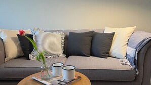 A grey sofa with striped and solid pillows, a small table with a magazine, two white mugs, and a vase with a single red rose.