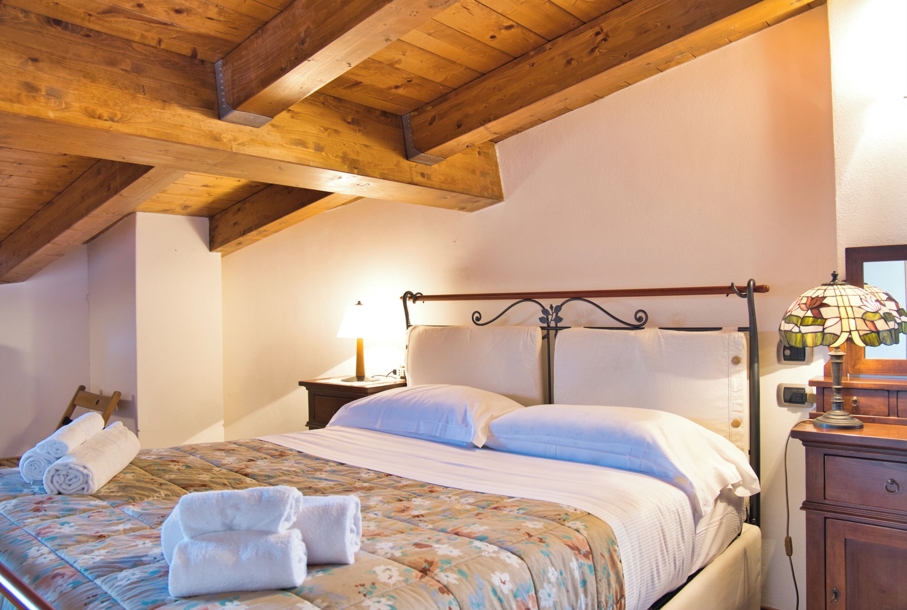 Un dormitorio con techo de madera, una cama con ropa de cama blanca, una mesita de noche con una lámpara y un aparador de madera.