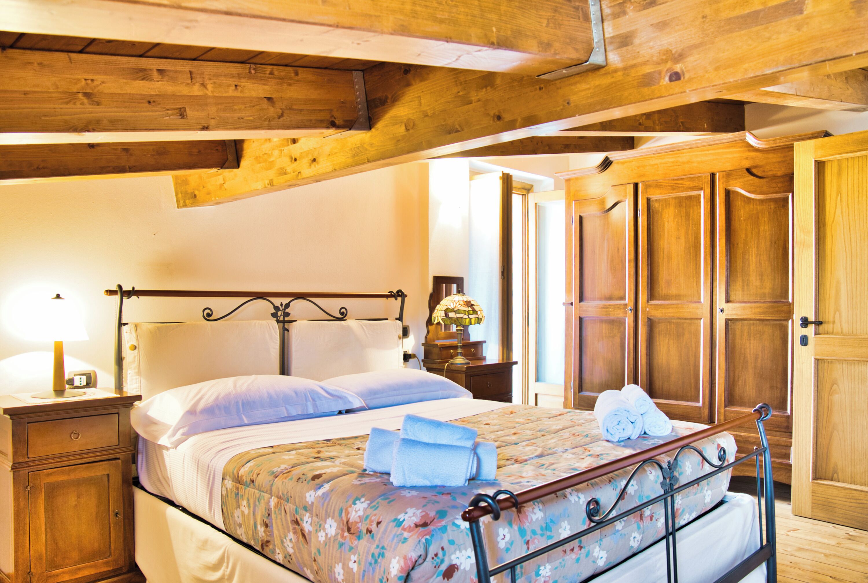 A bedroom with a wooden ceiling, a bed with a floral bedspread, a nightstand with a lamp, and wooden wardrobes.