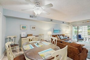 Une pièce comprenant une table à manger, des chaises, un canapé, un ventilateur de plafond, des œuvres d’art accrochées aux murs et une vue sur l’extérieur, visible à travers une porte vitrée.