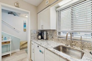 A kitchen with a granite countertop, stainless steel sink, white cabinets, and a window with blinds.