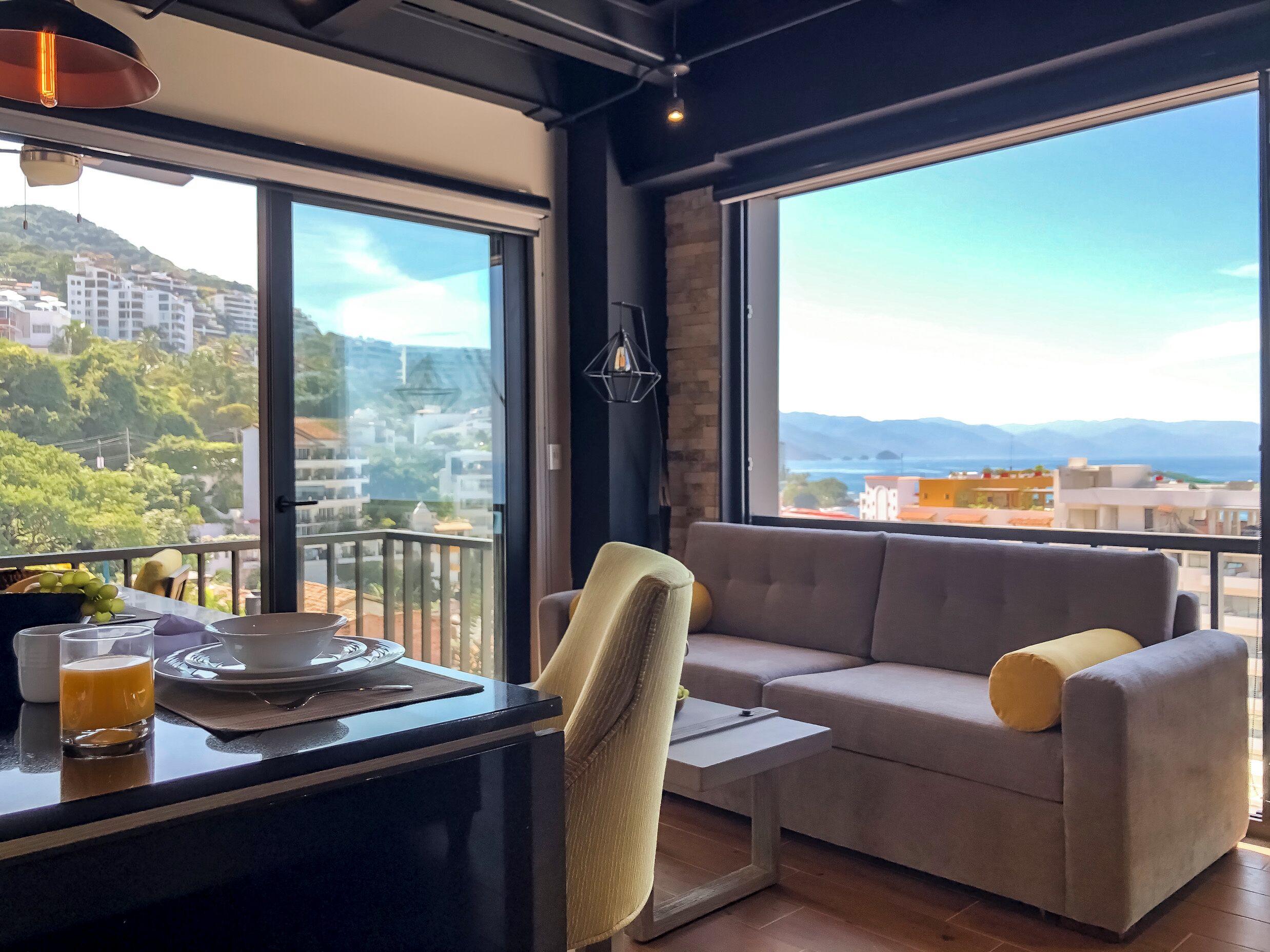Ein modernes Wohnzimmer mit einer Couch, einem kleinen Tisch und einem Glas Orangensaft. Ein großes Fenster bietet Blick auf Gebäude und Grünflächen.