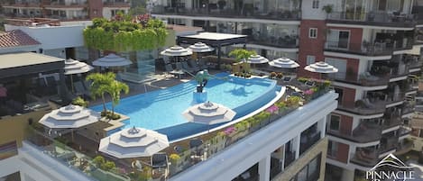 A rooftop pool with sun loungers and umbrellas, surrounded by balconies and greenery.