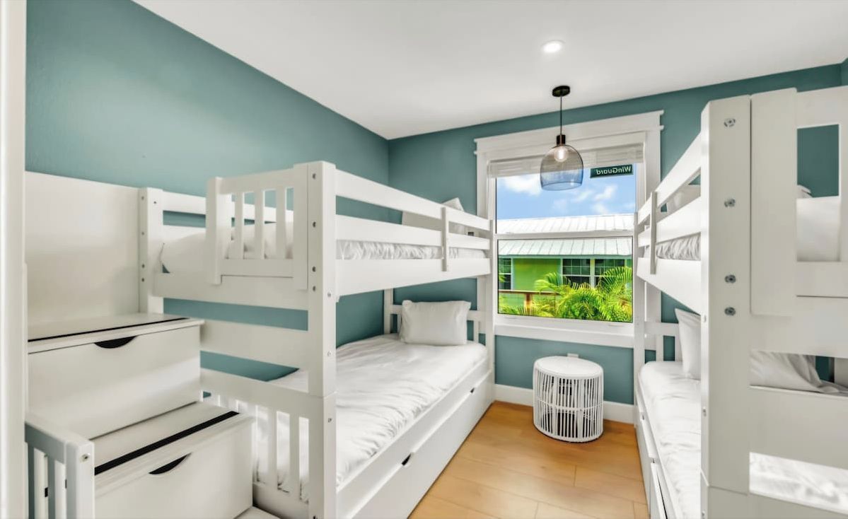 A room with bunk beds, a window with a view of greenery, and a white circular stool.