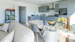 A modern kitchen with grey cabinets, a dining table with a floral tablecloth, and a sofa in the living area. - 6 Gwel Yr Wyddfa (Ty Croes)