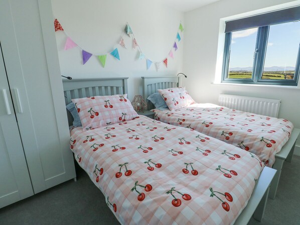 A bedroom with two beds, each with a pink and white checkered bedspread featuring cherries. There is a window with a view of the countryside, and a string of colorful triangular flags on the wall.