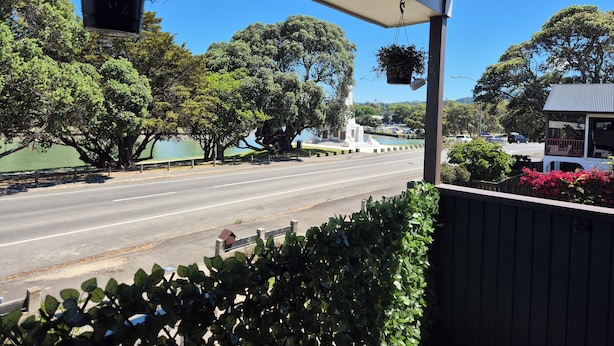 A view from a covered porch overlooking a road with trees and a body of water. - Esplanade Townhouse  (Gisborne)