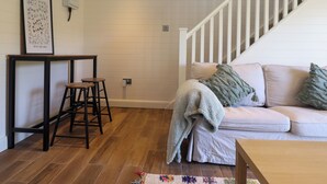 Ein Wohnzimmer mit Holzboden, einem weißen Sofa mit Kissen, einem kleinen Tisch mit Hockern, einer Treppe und einer Wandleuchte.