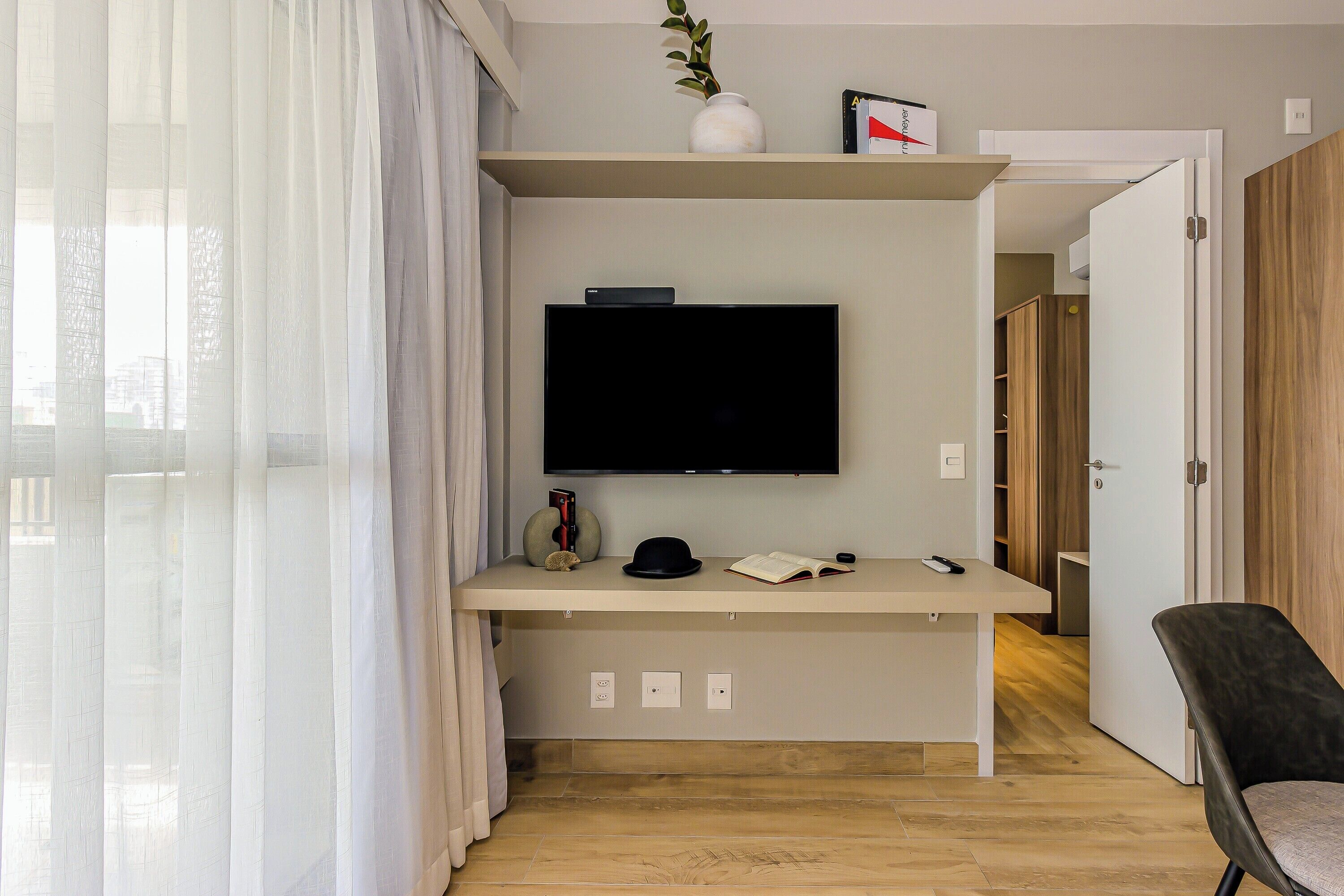 A modern room with a flat-screen TV mounted on a wall, a desk with a remote control, a hat, and a book.