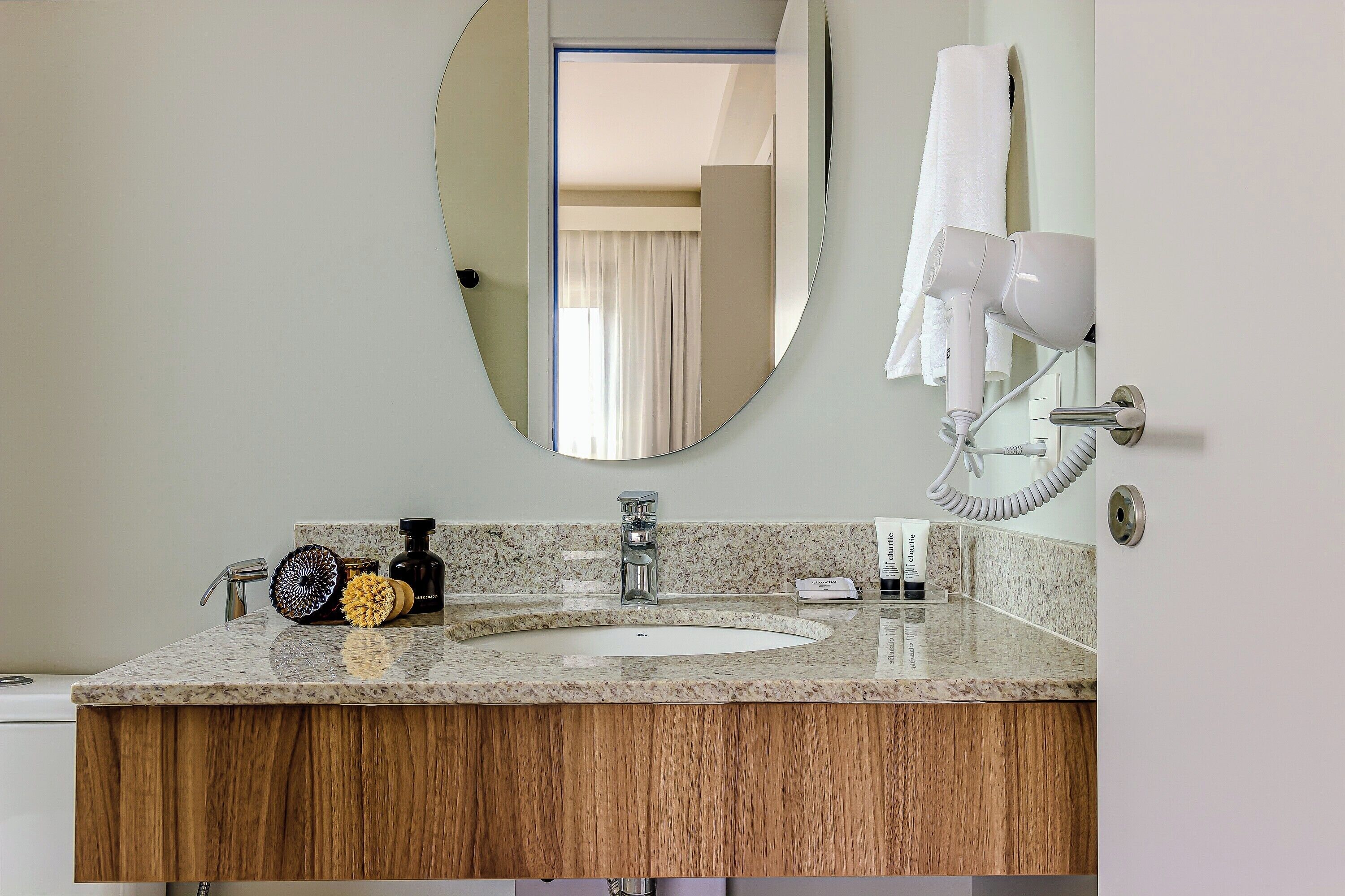 A bathroom with a granite countertop, a wooden vanity, a round mirror, a hairdryer, and a towel hanging on the wall.